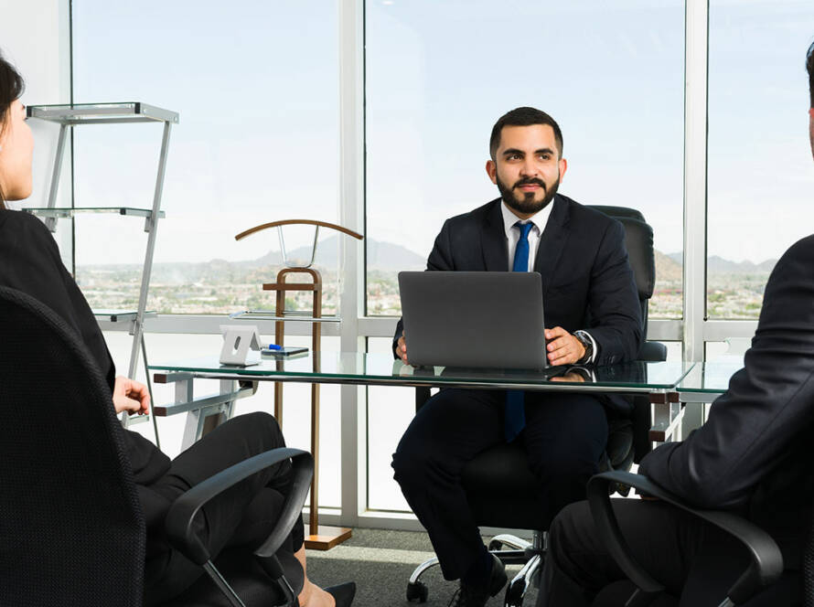 Executive behind glass desk in corner office out-looking city skyline while talking to a man and woman sitting across the table in business attire.