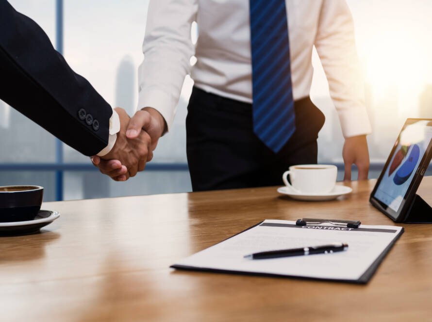 Two businessmen shaking hands over a contract at a desk overlooking city skyscrapers with tablet computer and coffee cup on desk.