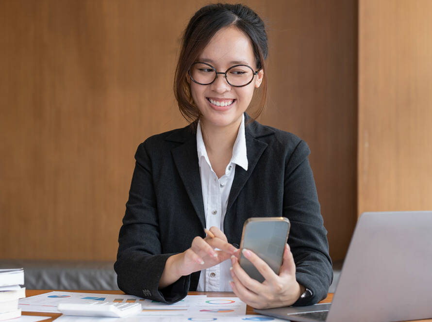 Asian woman smiling while typing on a phone in front of a laptop while at work.