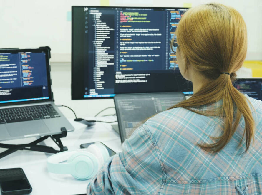 Woman with ponytail working in front of a large monitor and laptop, coding away.